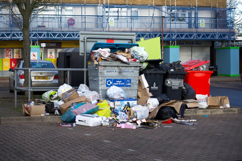 Two large black plastic bags of rubbish are placed on the pavement beside a curb, leaning against a metal fence with vertical bars. The bags appear to contain household waste, with visible crumpled pl